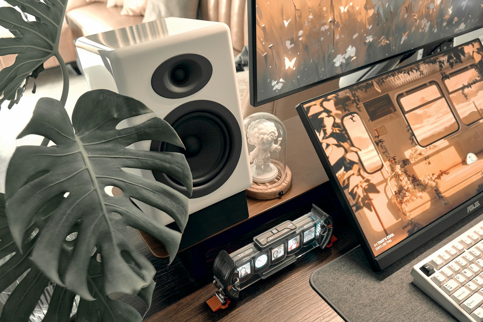 A monstera plant beside a white speaker, two monitors and a keyboard on a wooden desk, a workspace for checking ad copy before launch.